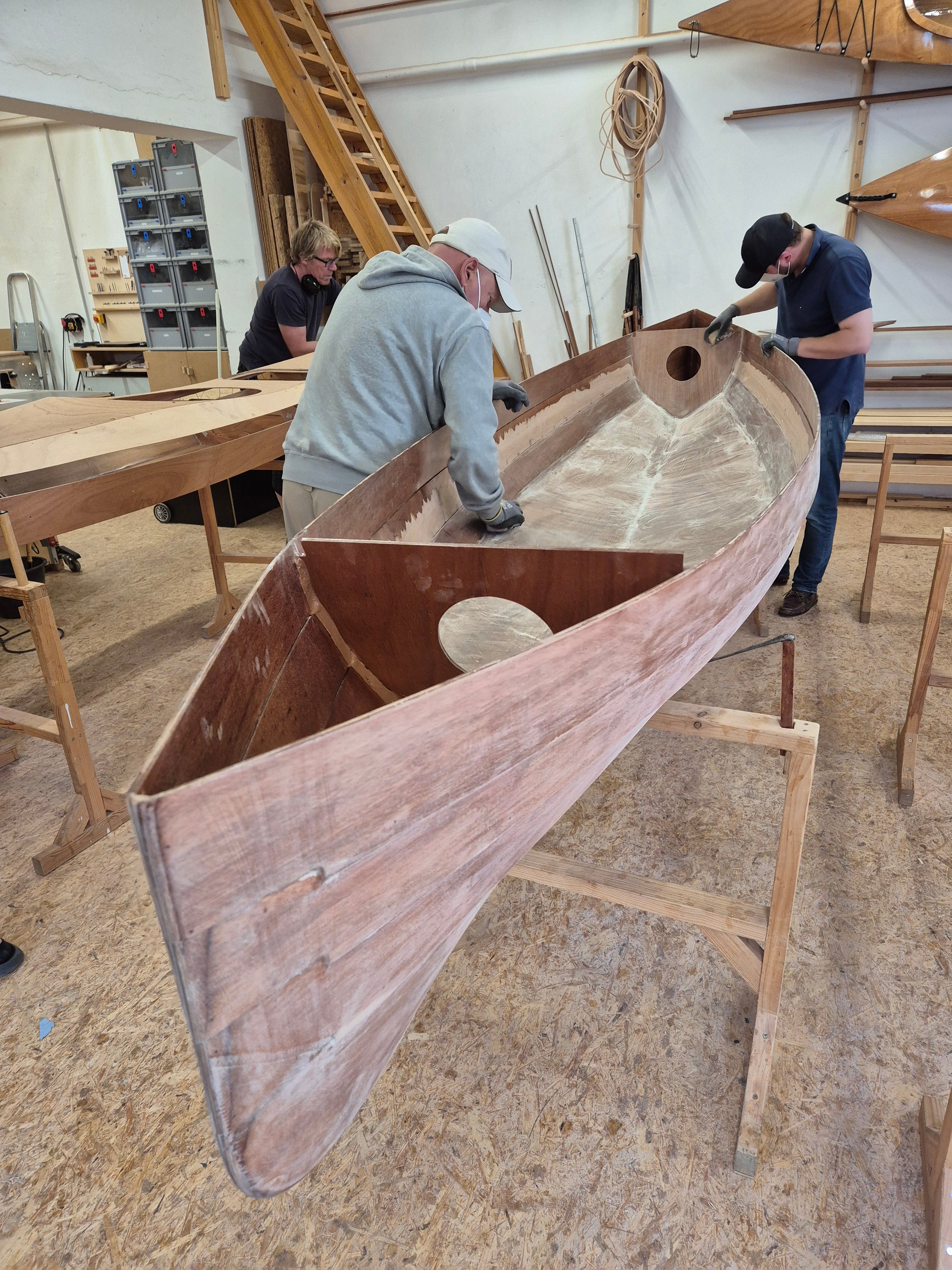Person schleift den Holzrumpf eines selbstgebauten Kanadiers in einer Werkstatt.